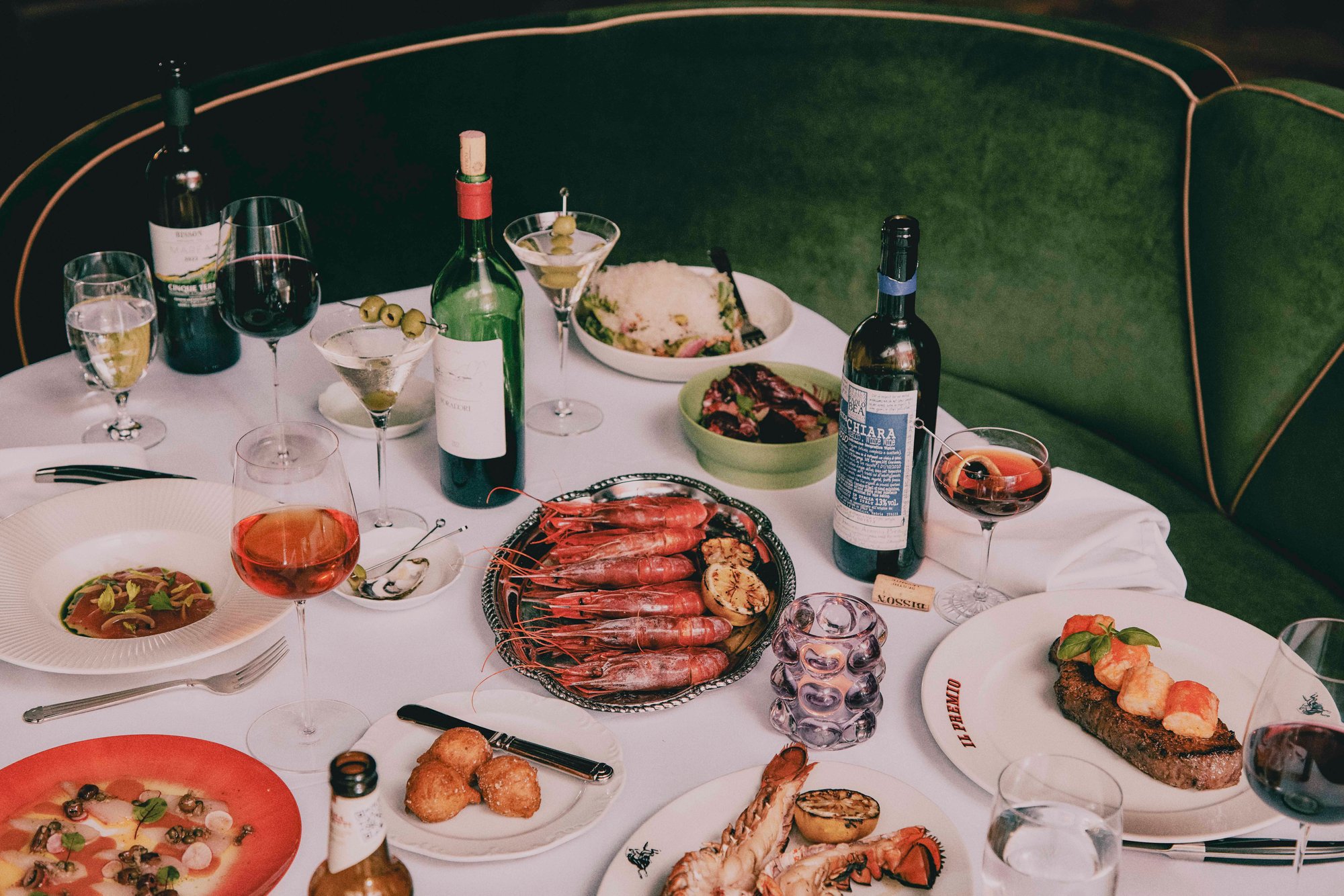 A table at Il Premio laid with wine, grilled prawns and pasta beside a green velvet banquette